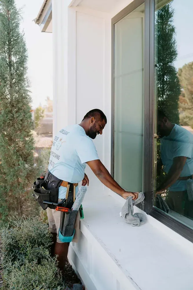 Worker sanding a white exterior windowsill beside a large glass window