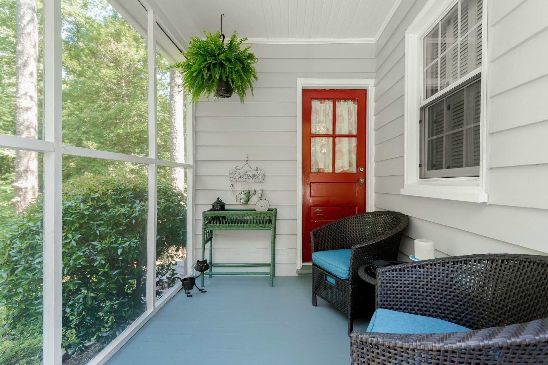 Screened porch with black wicker chairs, hanging fern, red door, and potted table by large windows