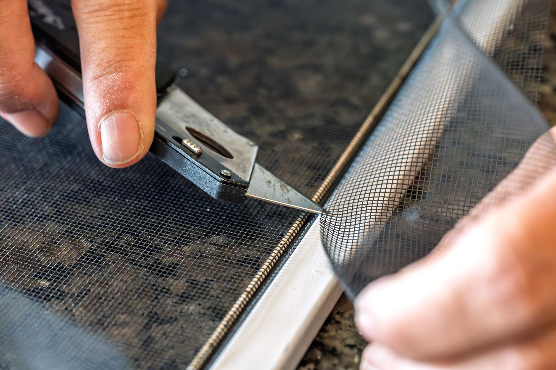 Hands using a utility knife to trim a black mesh screen on a frame.