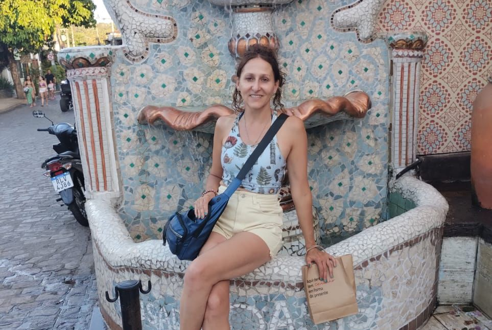 Una persona está sentada en el borde de una fuente ornamentada con mosaicos en una calle, vestida con una blusa estampada.