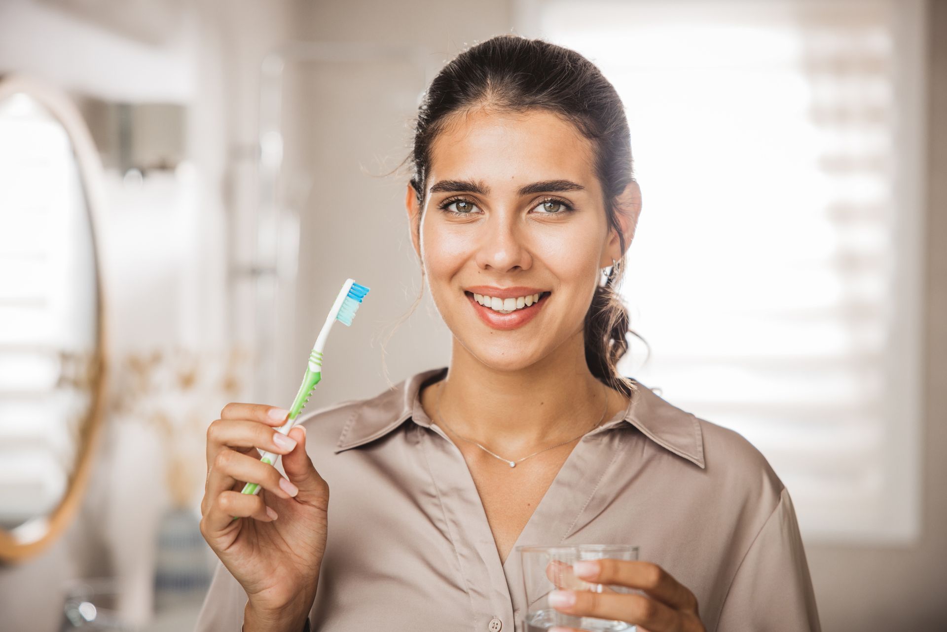 Smiling person holding a toothbrush and glass of water in a clean bathroom.