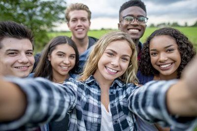 Five smiling friends taking a selfie outdoors in front of greenery