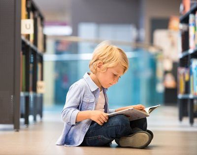 Child sitting on a library floor, reading an open book between bookshelves.