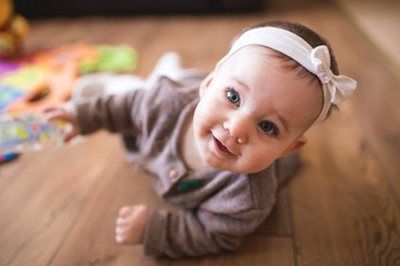 Baby crawling on a wooden floor, looking up with a white headband and gray outfit.