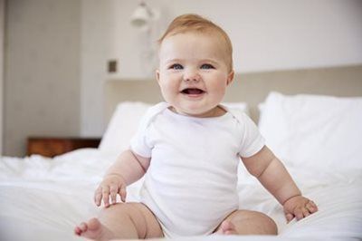 Smiling baby in a white onesie sitting on a bed in a bright bedroom