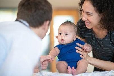 Adult helping a baby sit while another adult smiles nearby in a bright indoor setting