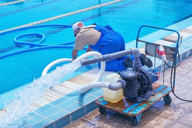 Worker operating a pump by a blue swimming pool, with water being drained onto the deck