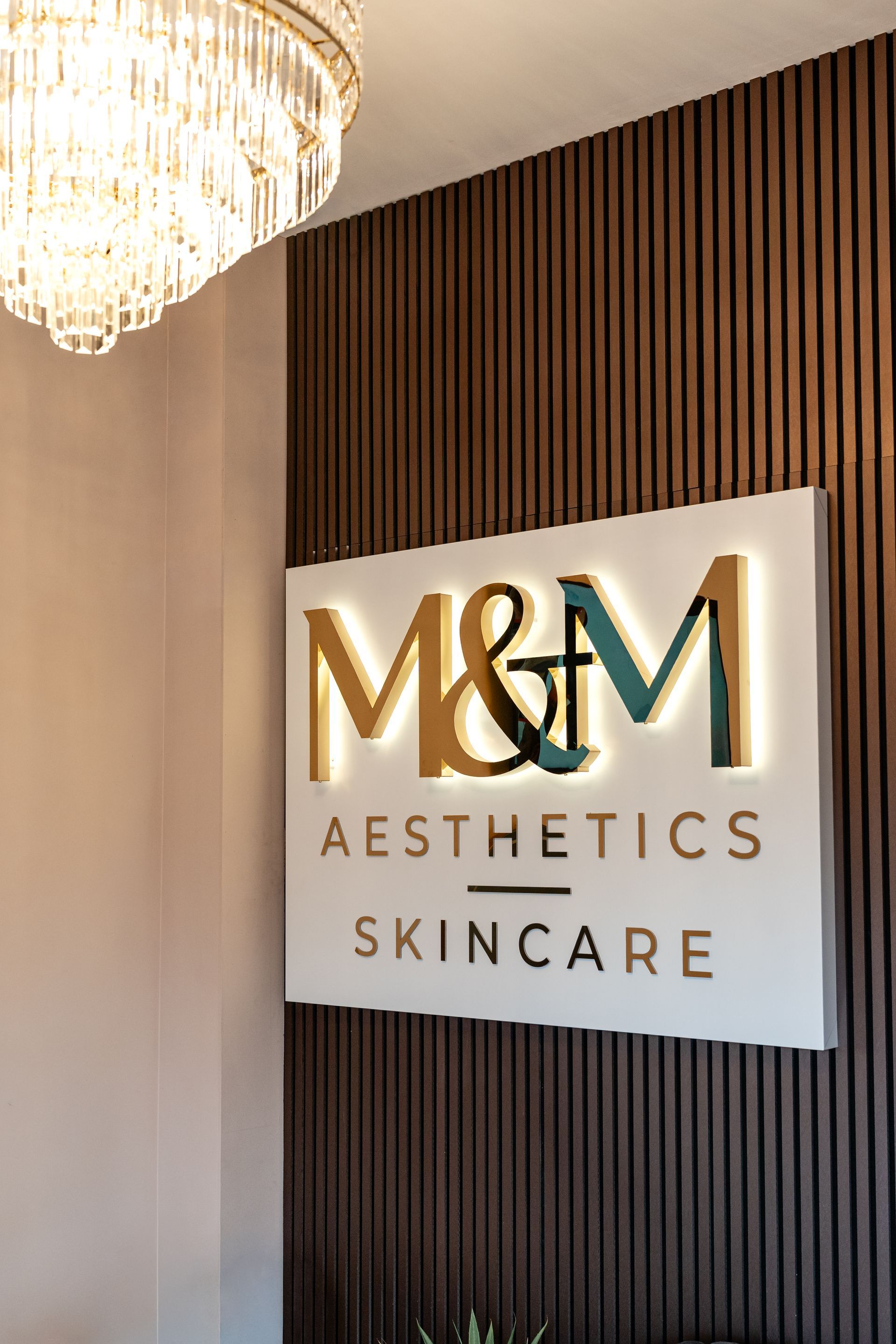 M&M Aesthetics Skincare sign on a wood-paneled wall beside a chandelier
