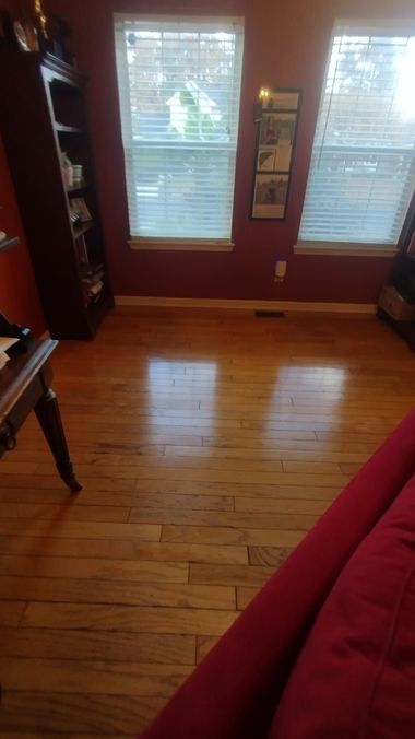 Empty living room with hardwood floor, two windows with blinds, and a red sofa edge in the foreground