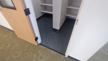 Open closet with white shelves and dark flooring beside a light wood door