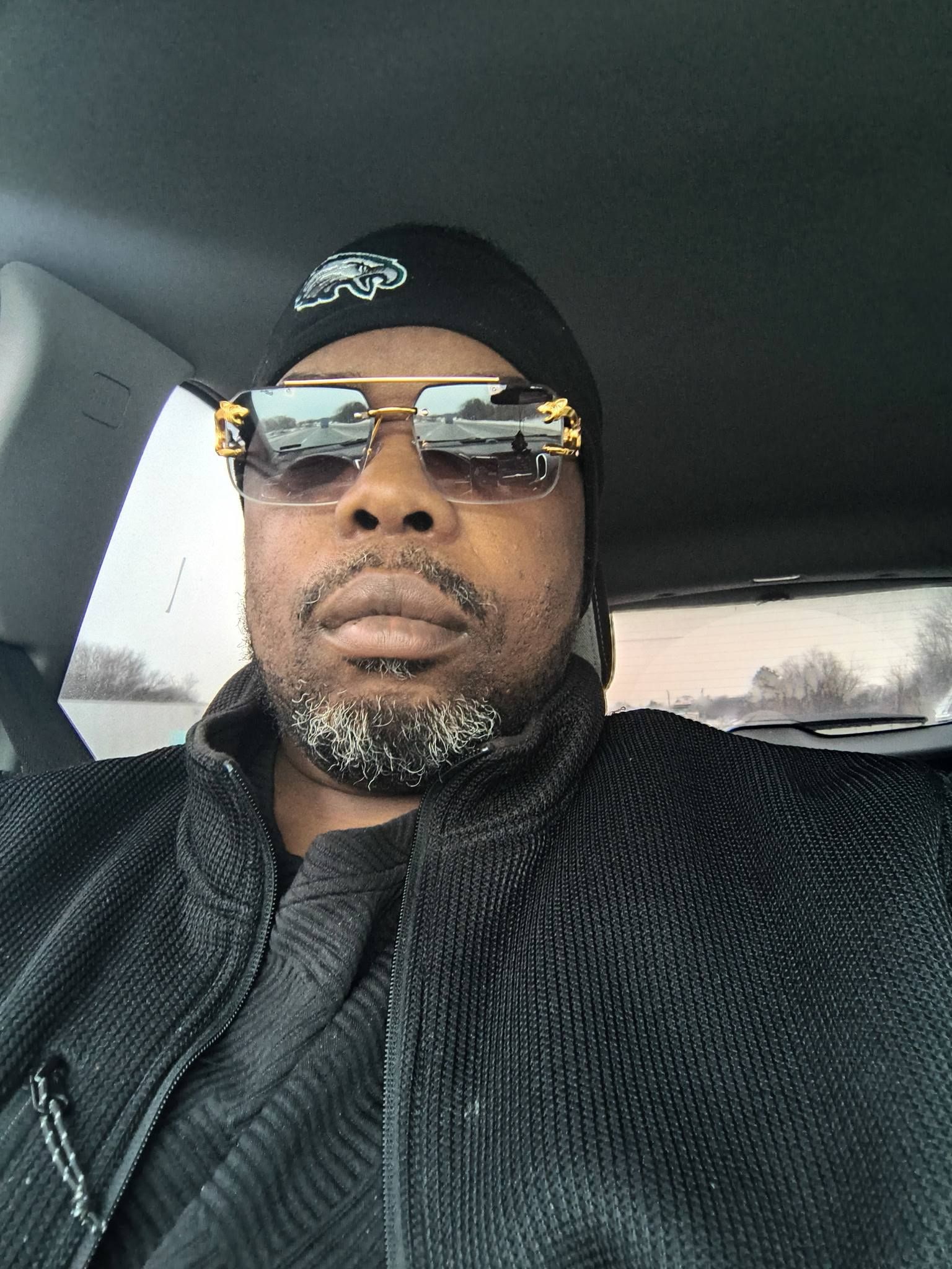 Man wearing sunglasses and a black beanie, seated in a car with a calm expression.