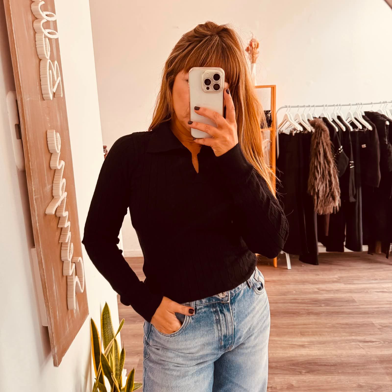Una persona se toma una selfie frente al espejo en una tienda de ropa, vistiendo una camiseta negra y jeans.
