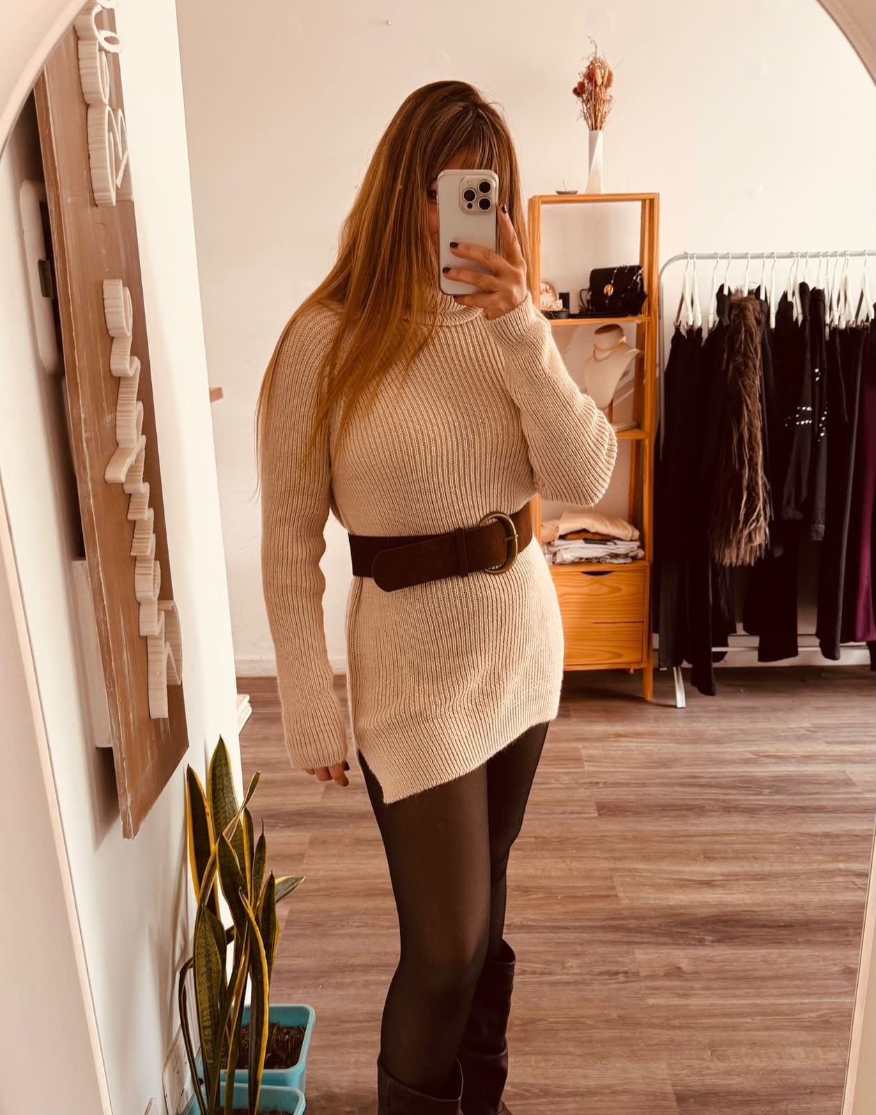Una persona se toma una selfie frente al espejo en una boutique, vistiendo un vestido de punto beige, medias negras y botas.