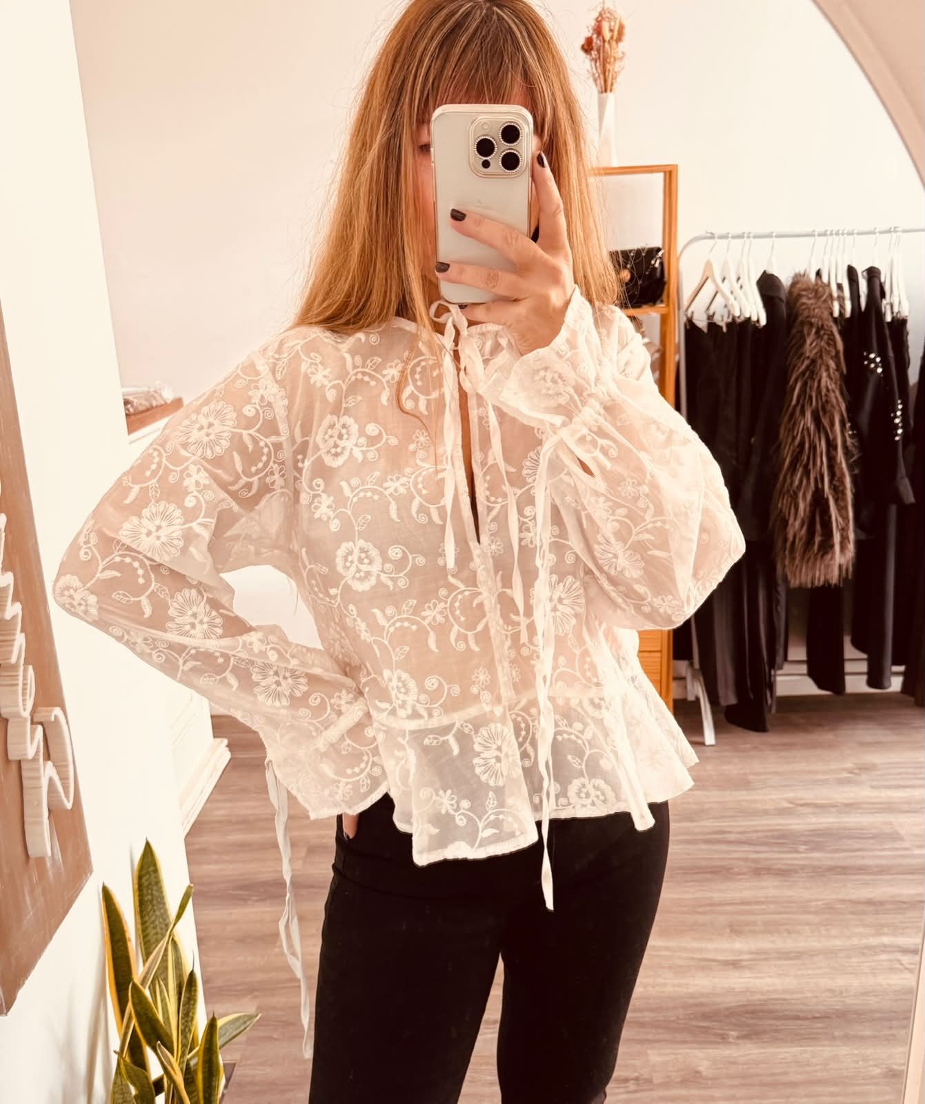 Una persona con una blusa blanca de encaje y pantalones negros se toma una selfie frente al espejo en una boutique de ropa.