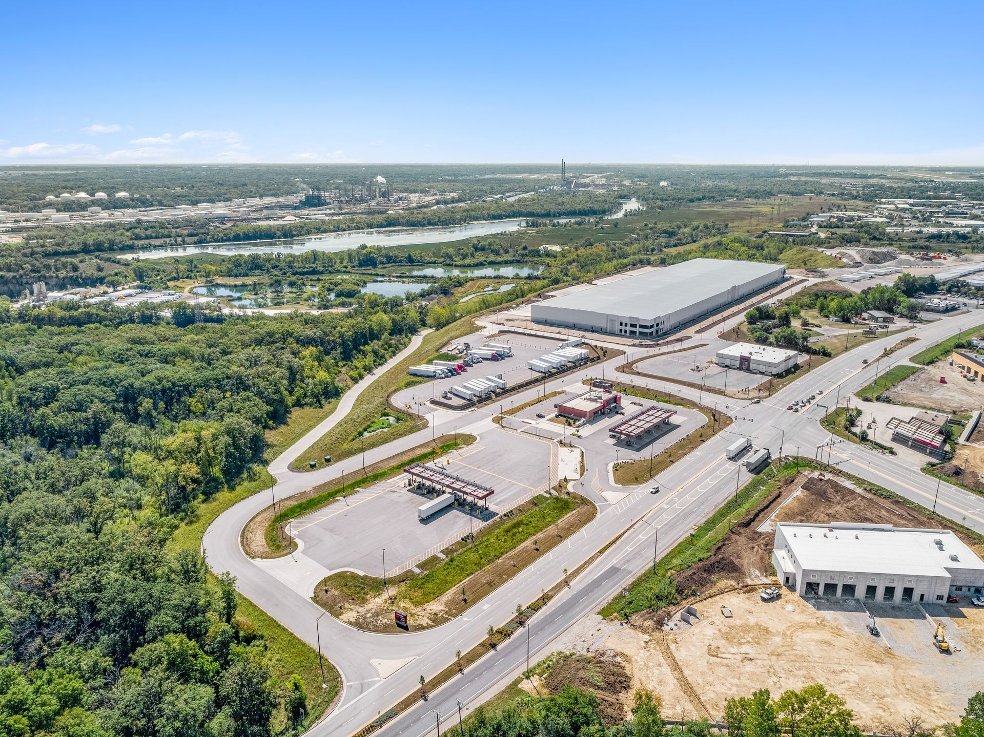 Aerial view of a large warehouse, several trucks, and other buildings surrounded by trees and roads under a blue sky.