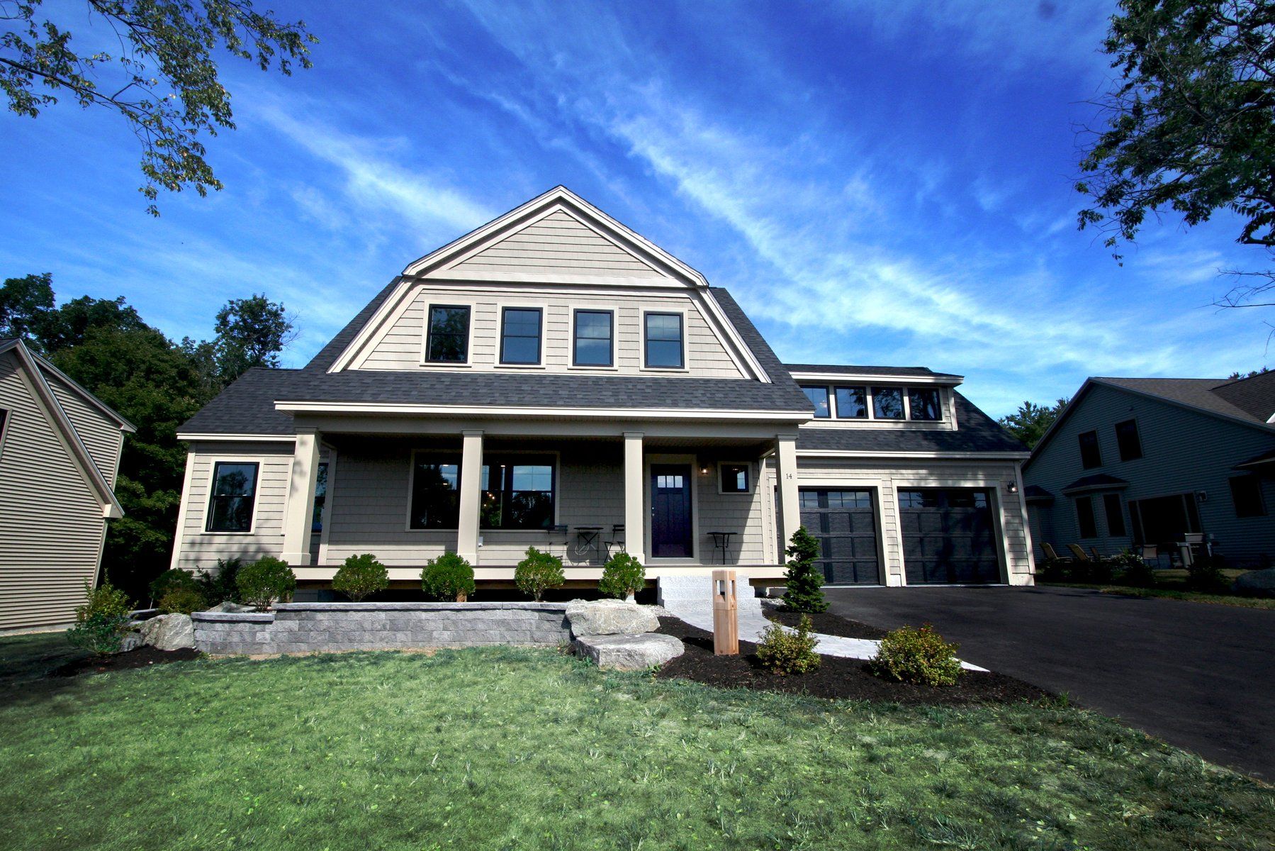 Exterior view of two story beige home with driveway that meets two garages, and large front yard with green grass and pumpkins, hay stacks, and yellow flowers for Fall season decor..