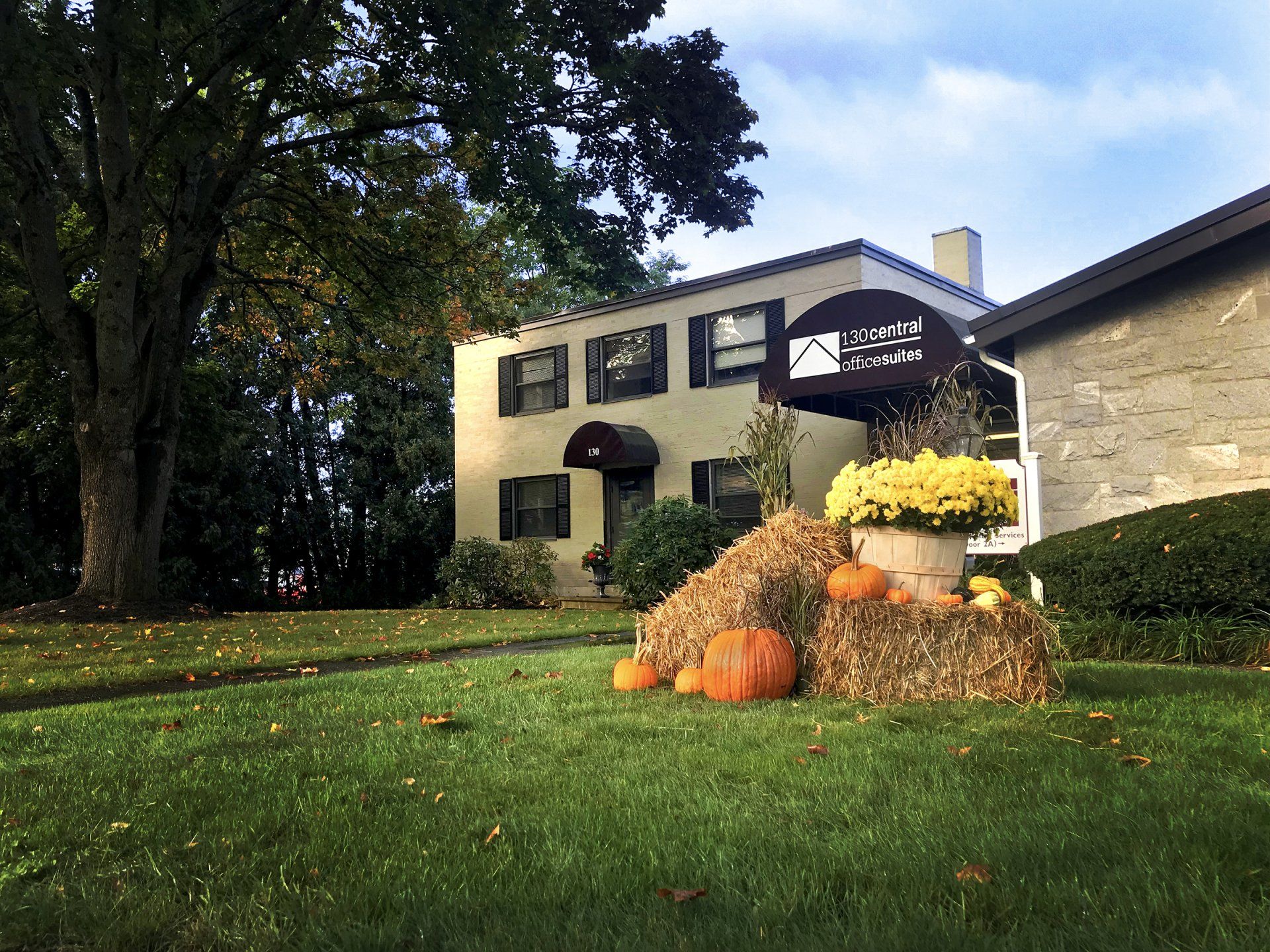 Exterior view of two story beige building of office suites with chimney, and large front yard with green grass and pumpkins, hay stacks, and yellow flowers for Fall season decor.