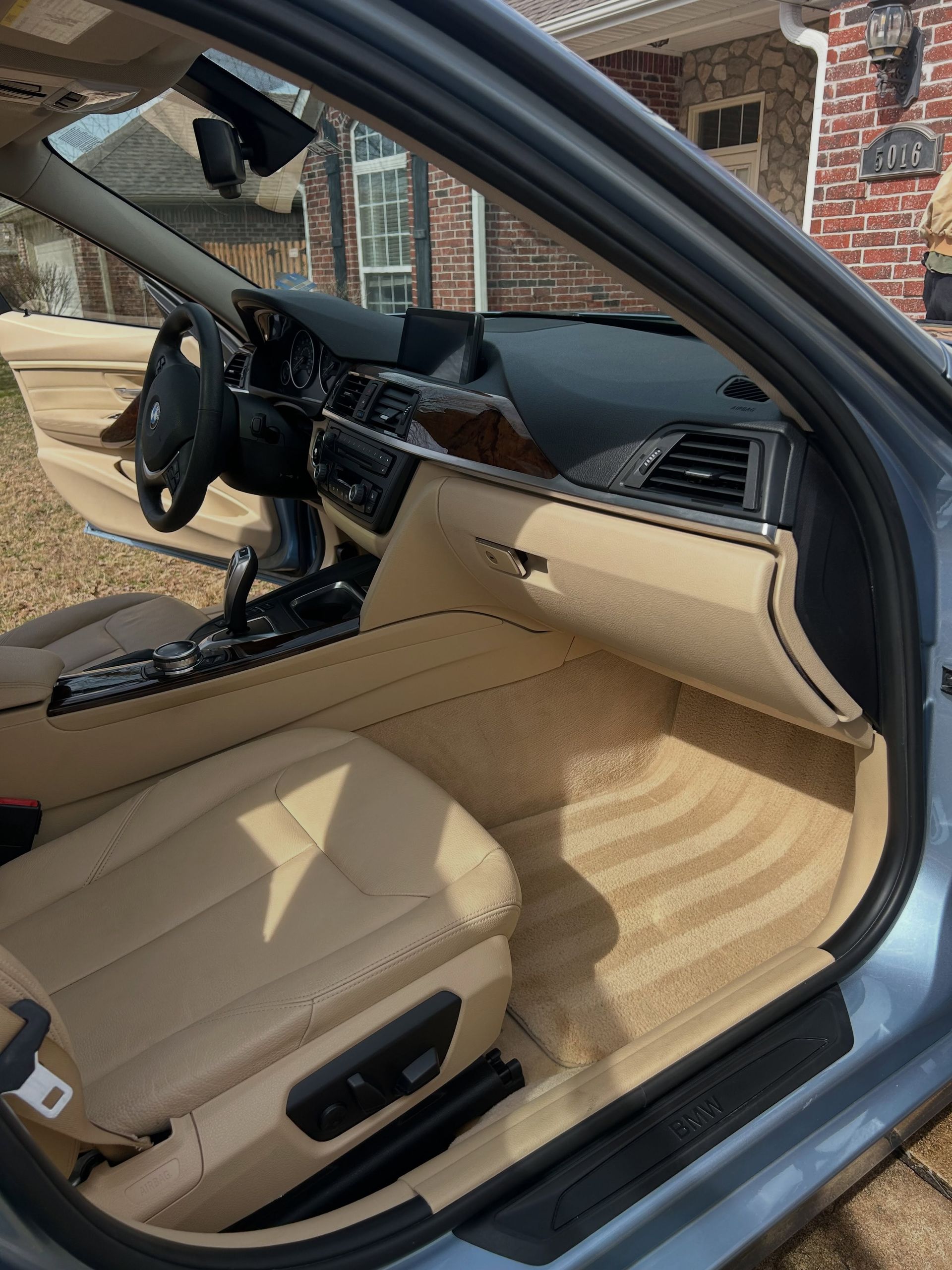 Car interior with tan leather seats, black steering wheel, and open door parked by a brick house.