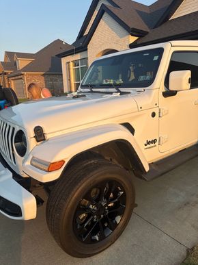 White Jeep Wrangler parked in a suburban driveway at sunset, with black wheels and a brick house behind it