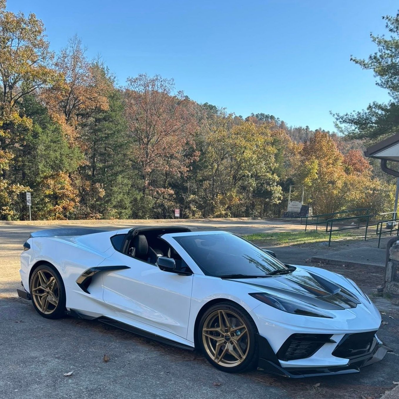 White sports car with gold wheels parked on pavement, with autumn trees in the background