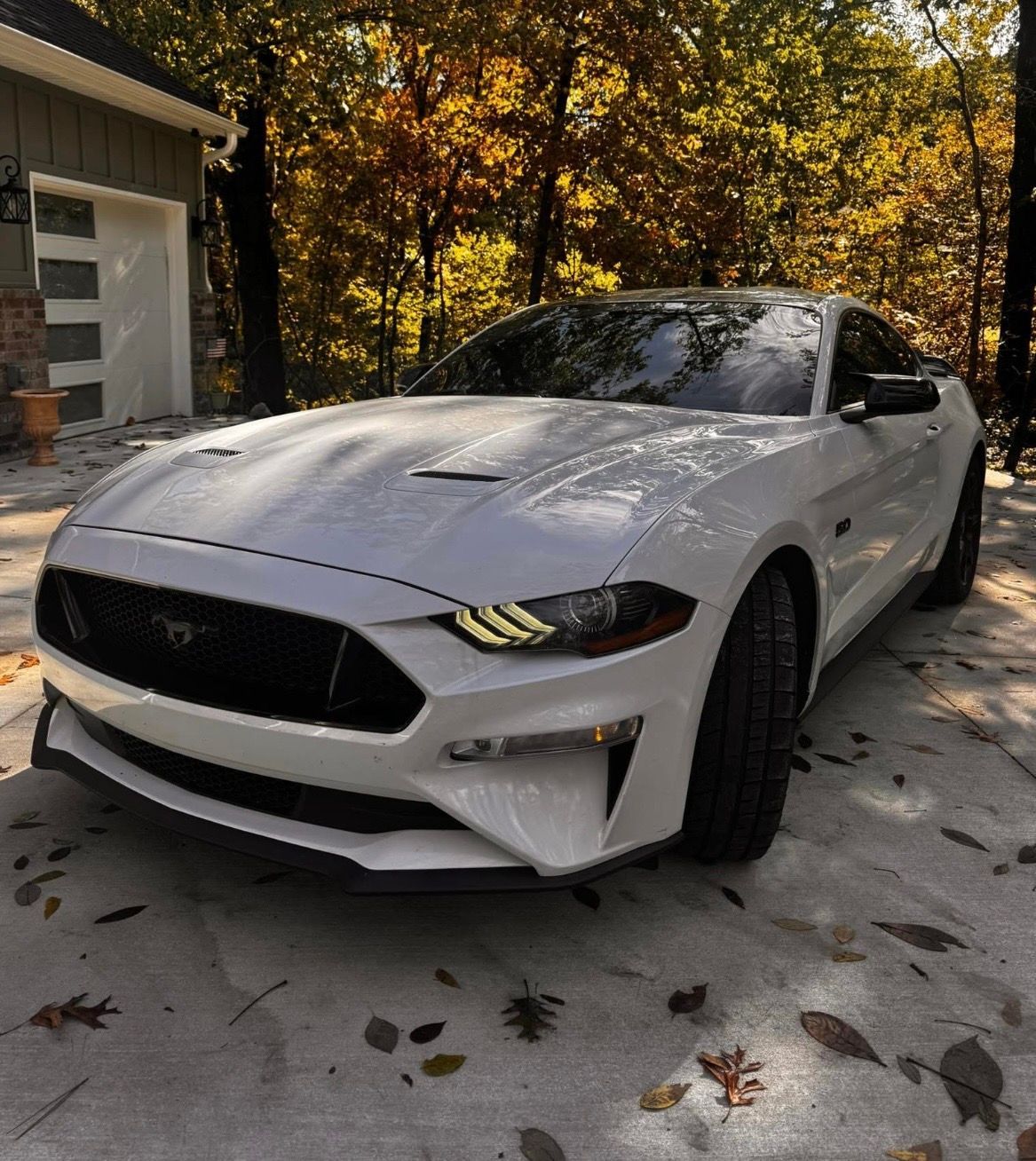 White sports car parked on a leaf-strewn driveway beside a garage in autumn trees