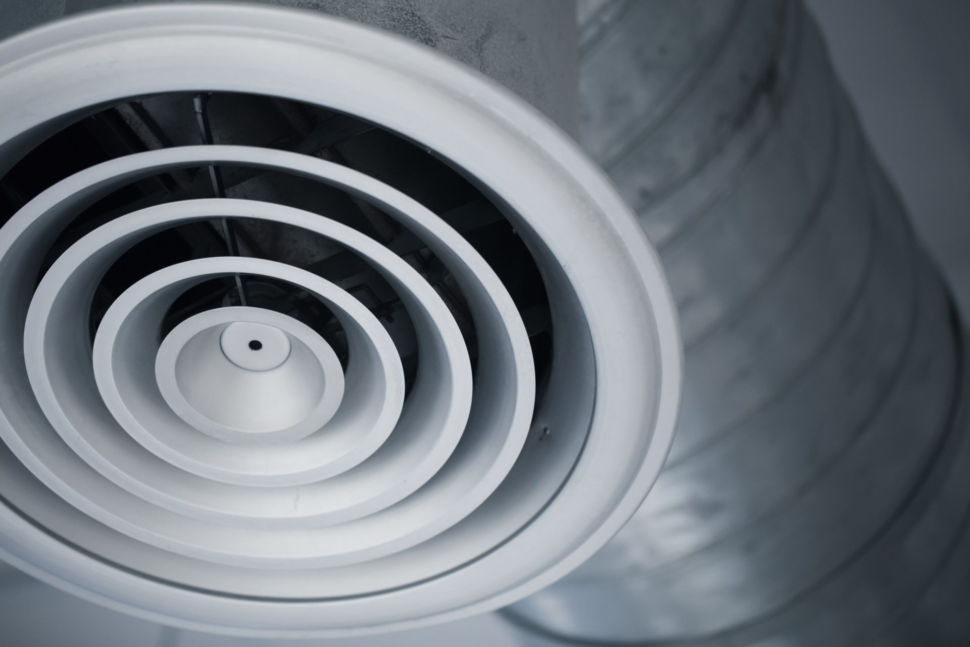 A white circular ceiling air diffuser vent connected to a silver metal industrial duct.