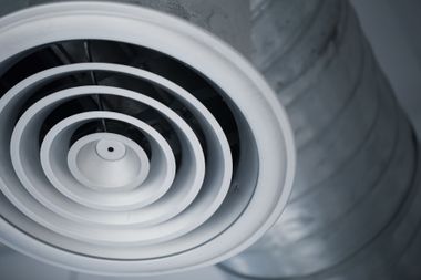 A white circular ceiling air diffuser vent connected to a silver metal industrial duct.