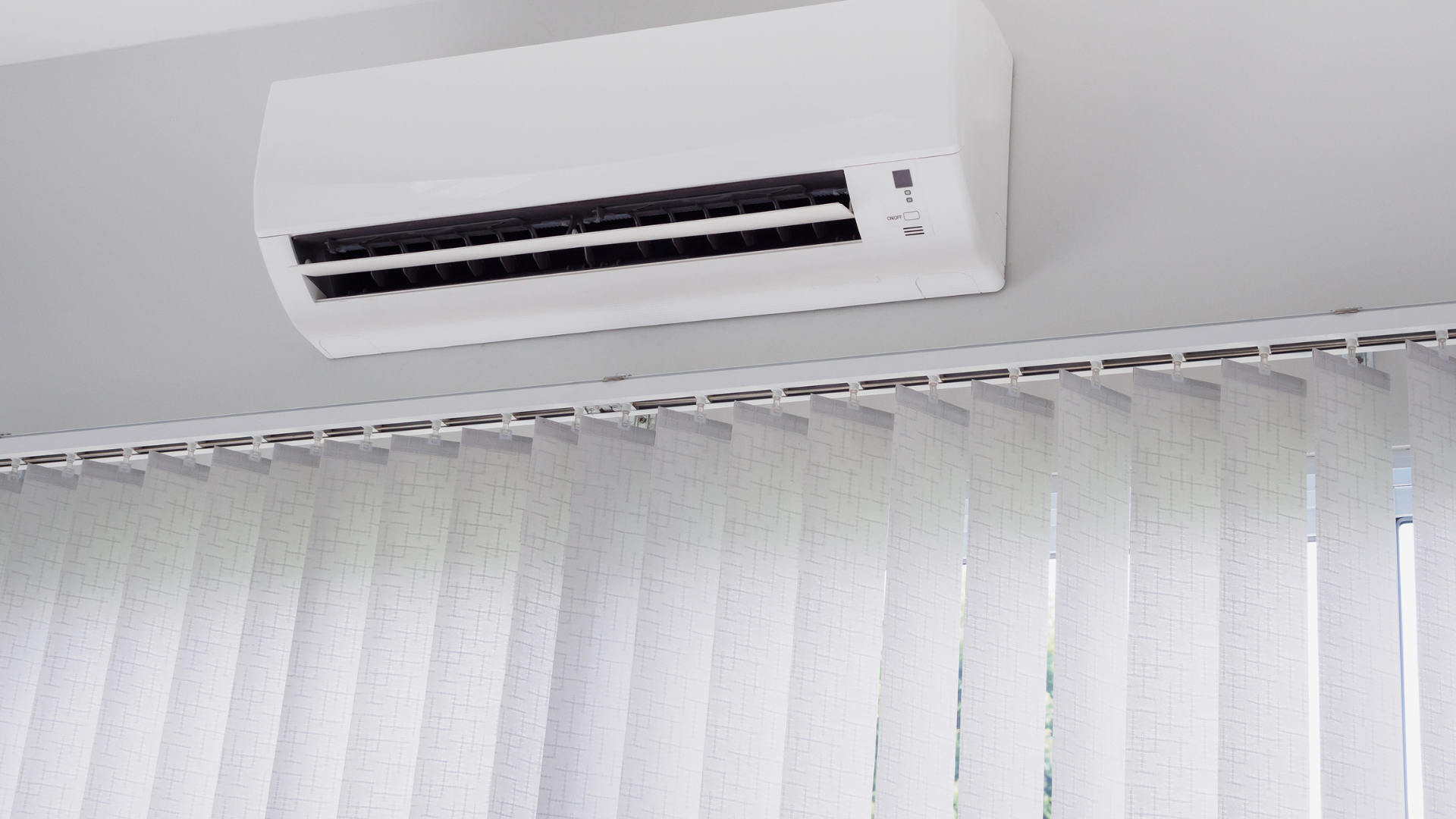 A white wall-mounted air conditioner positioned above a set of white vertical blinds.