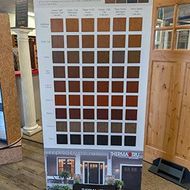 Display of brown exterior paint samples on a showroom board beside a wooden door, with house photos below