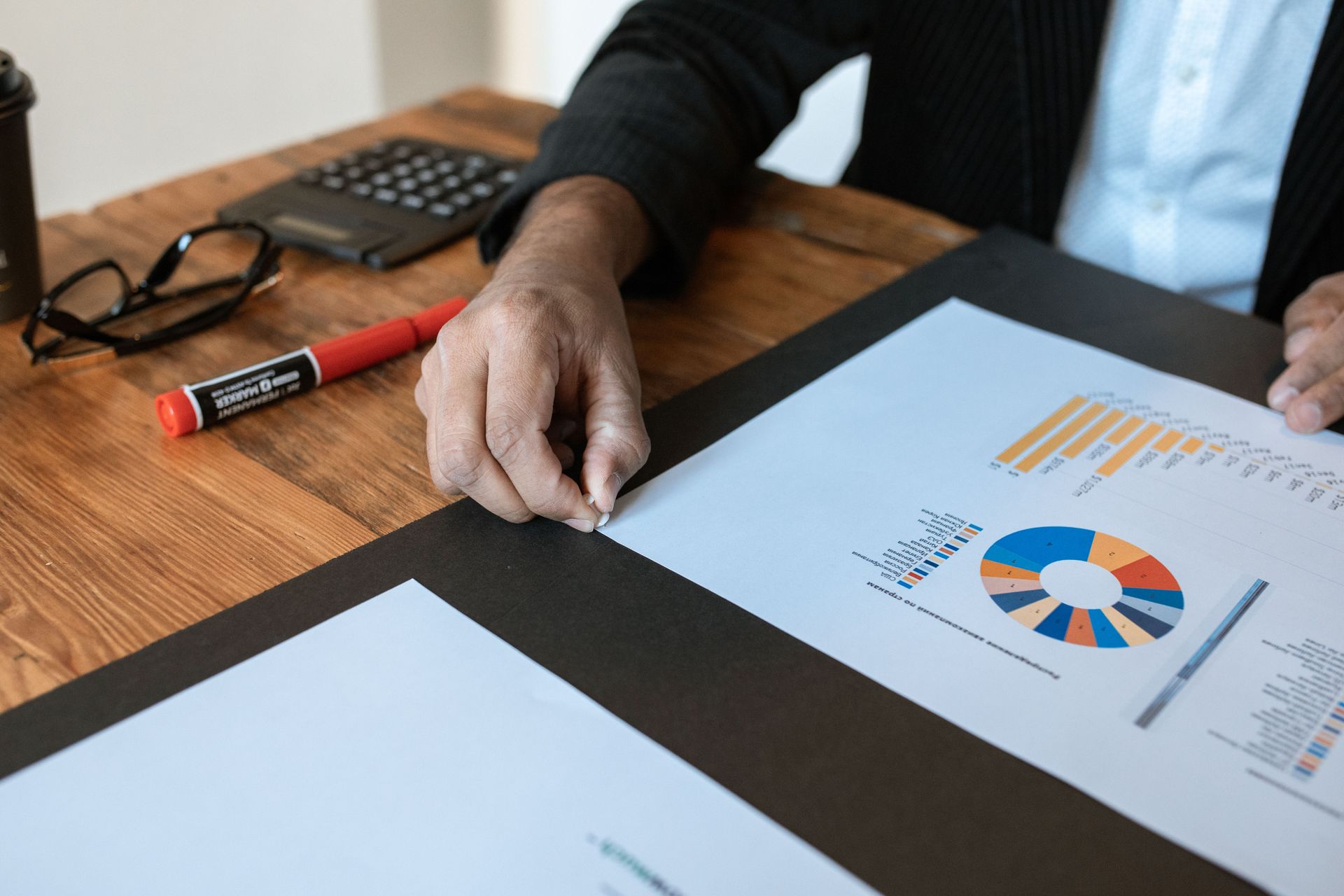 Person reviewing printed charts and graphs at a wooden desk with a laptop and marker nearby