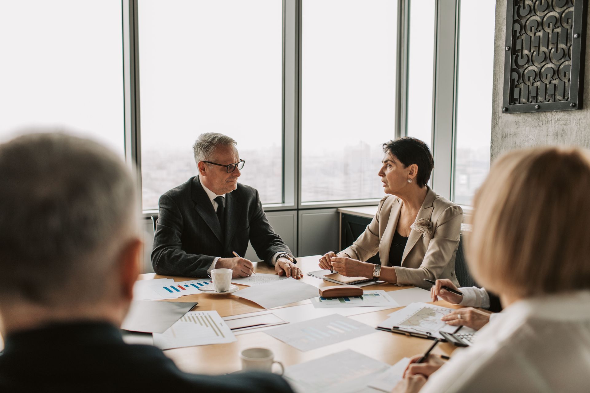 Business meeting around a table in a bright office, with documents spread out and two people speaking.