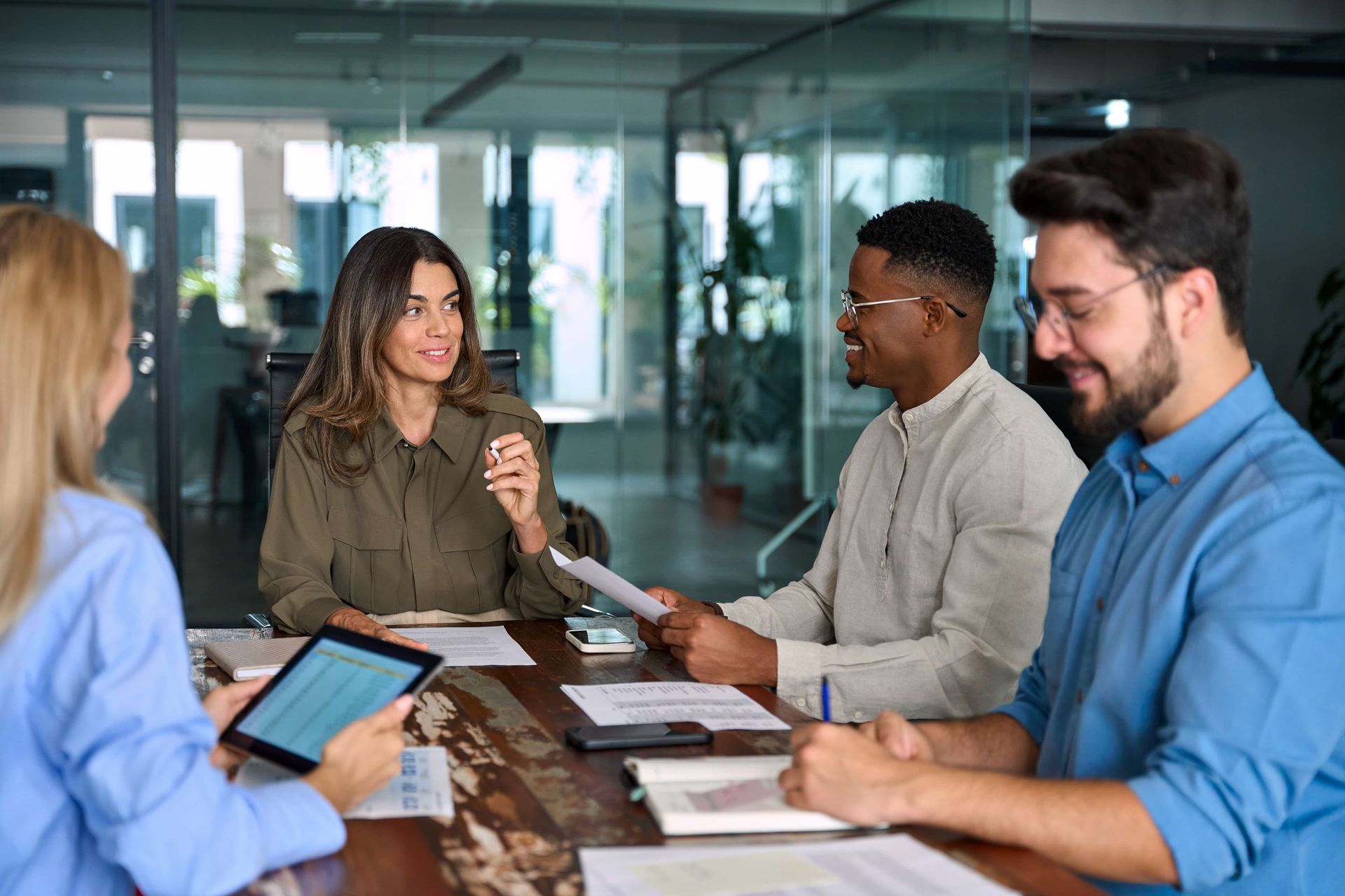 Colleagues in a glass-walled office meeting around a table with documents and a tablet
