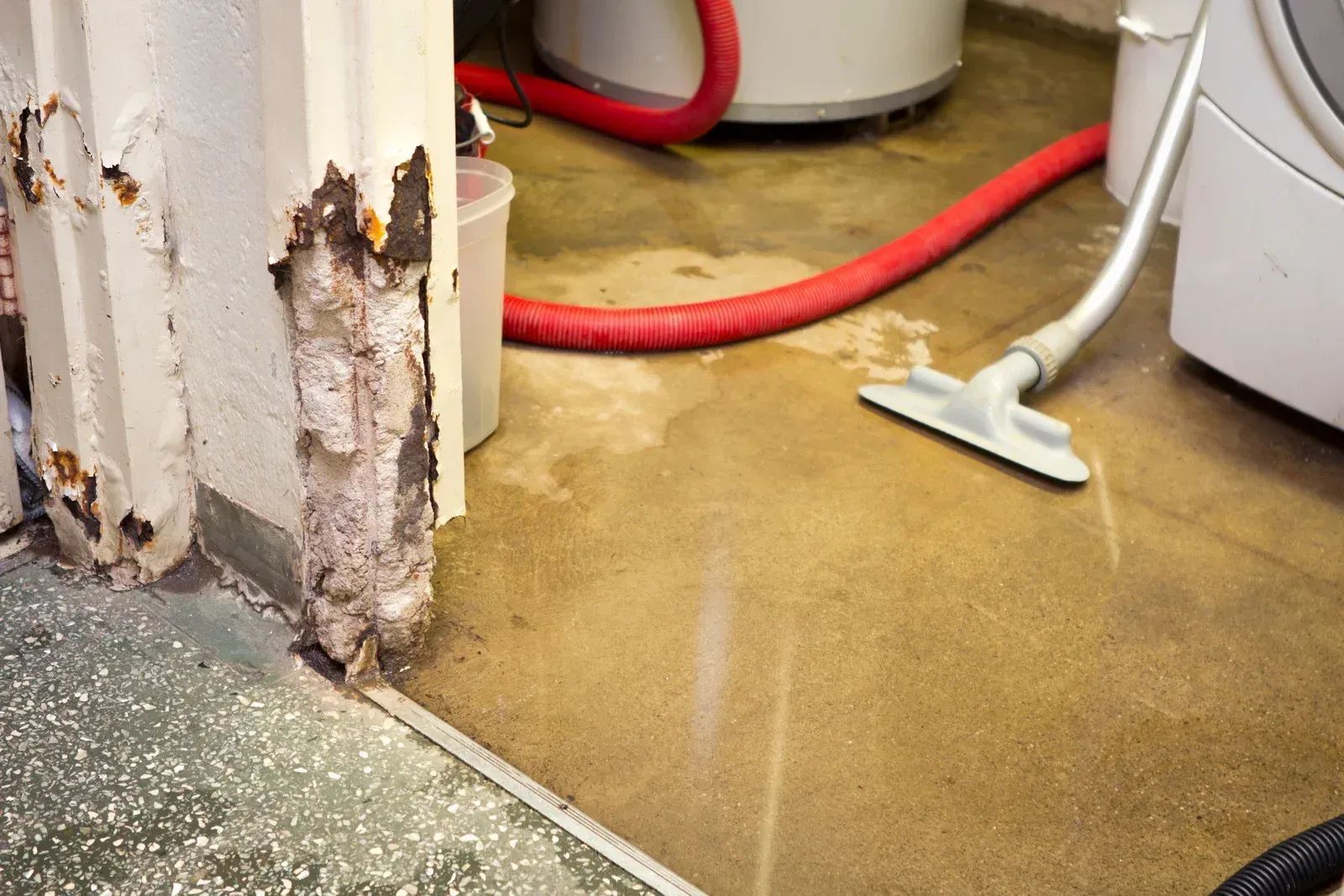 Damaged wall corner with peeling paint beside a red hose and mop on a wet floor