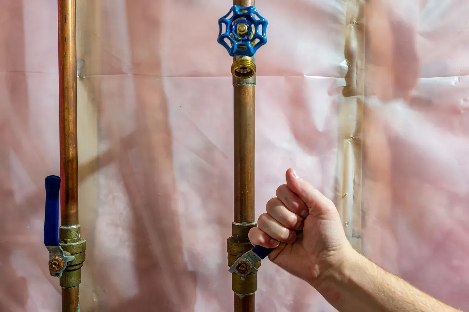 Hand turning a blue valve handle on a copper pipe against a pink background