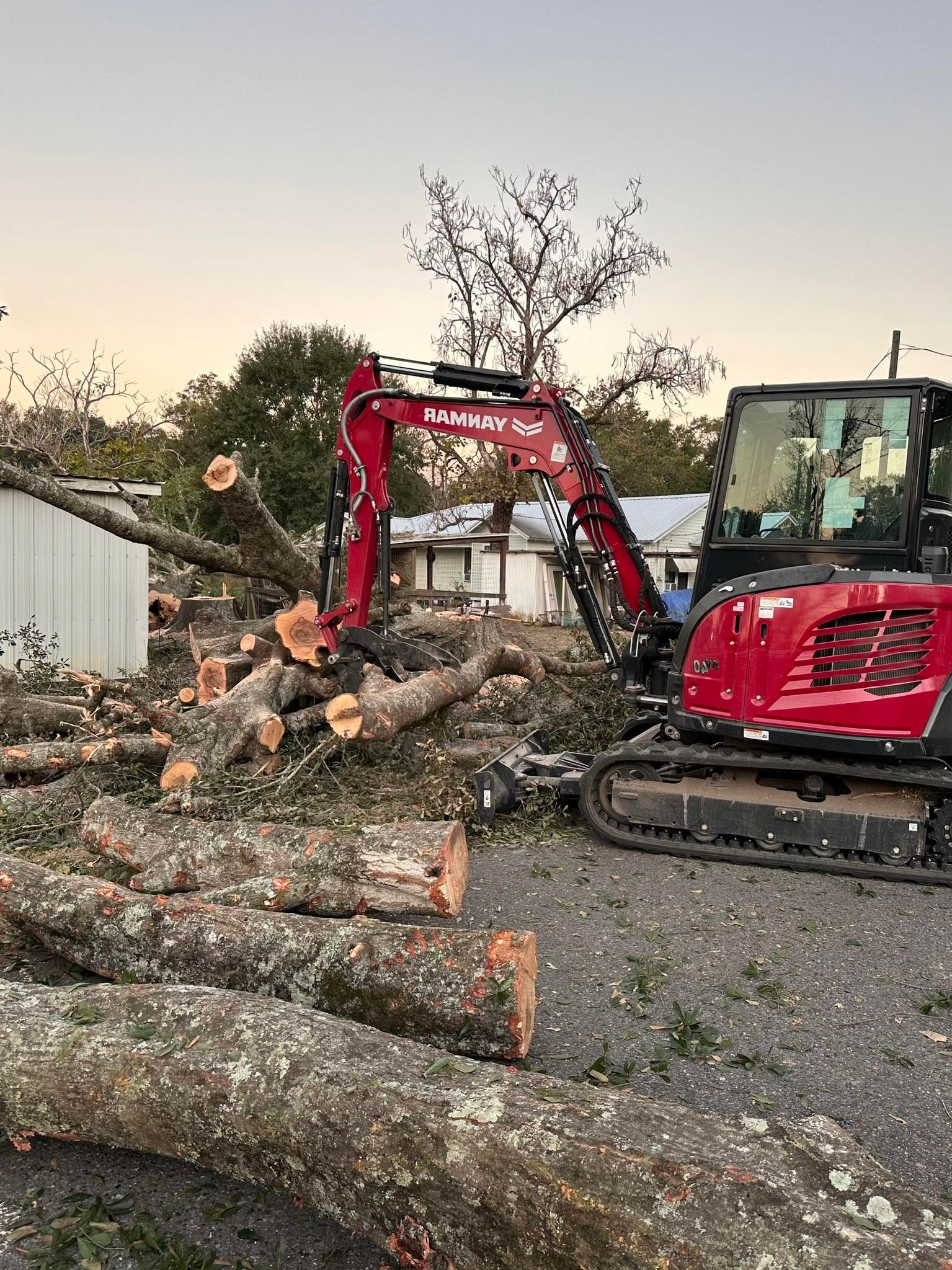 Red excavator lifting cut tree trunks at a gravel lot beside a white building at dusk