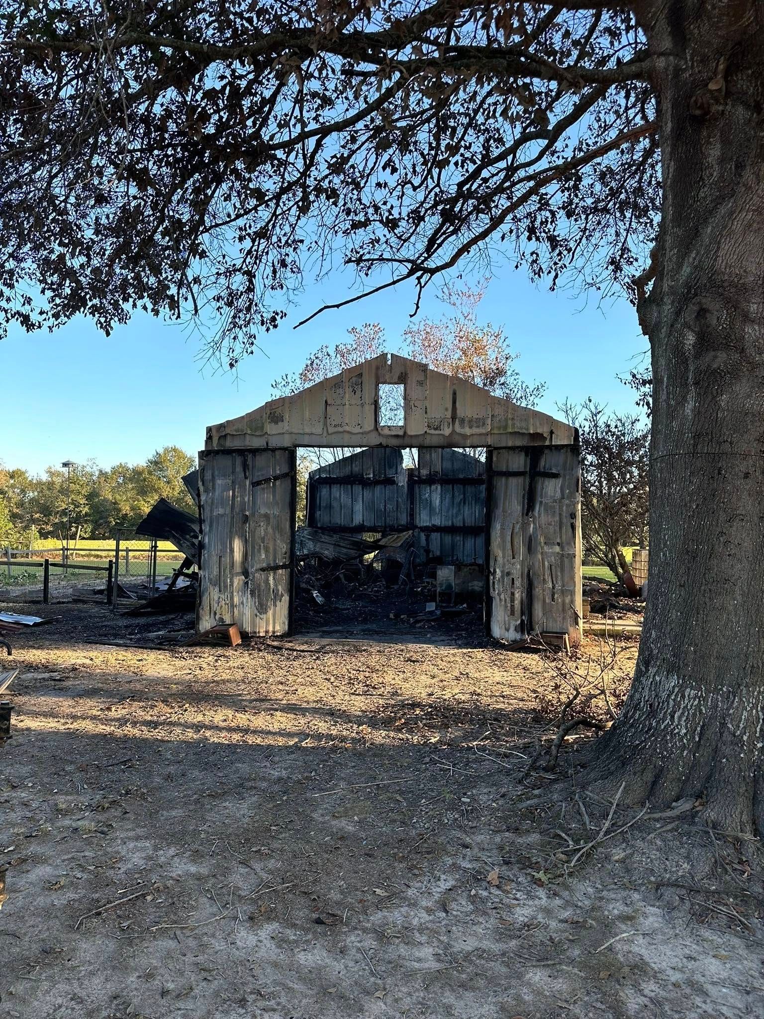 Old weathered shed with dark doors under a large tree in a dry, open field.
