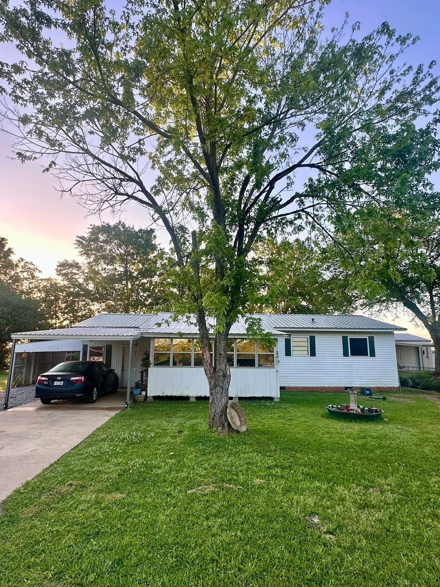 White house with a large tree in the front yard and a car parked under the carport at dusk