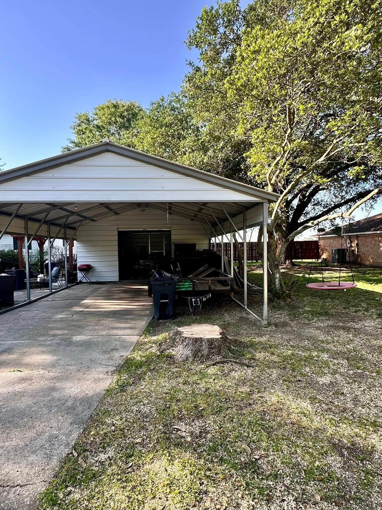 Carport beside a house with a gravel driveway, trash bins, and a large tree in the yard.