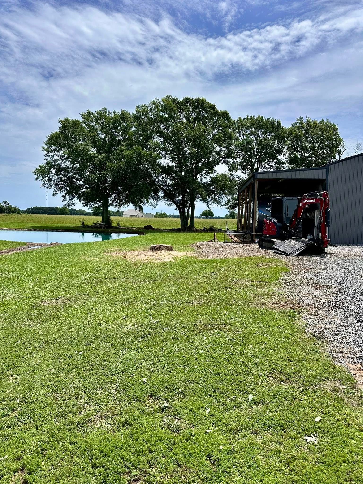 Gravel path beside a lake with two trees and a red mower under a shed on a sunny day