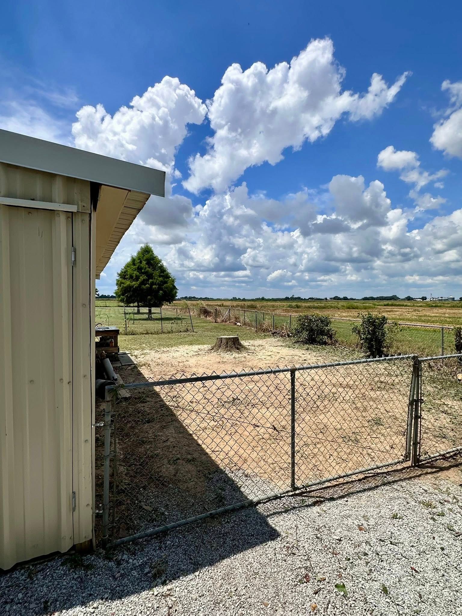 Metal shed beside a chain-link gate, opening to a dry field under a bright blue sky with clouds