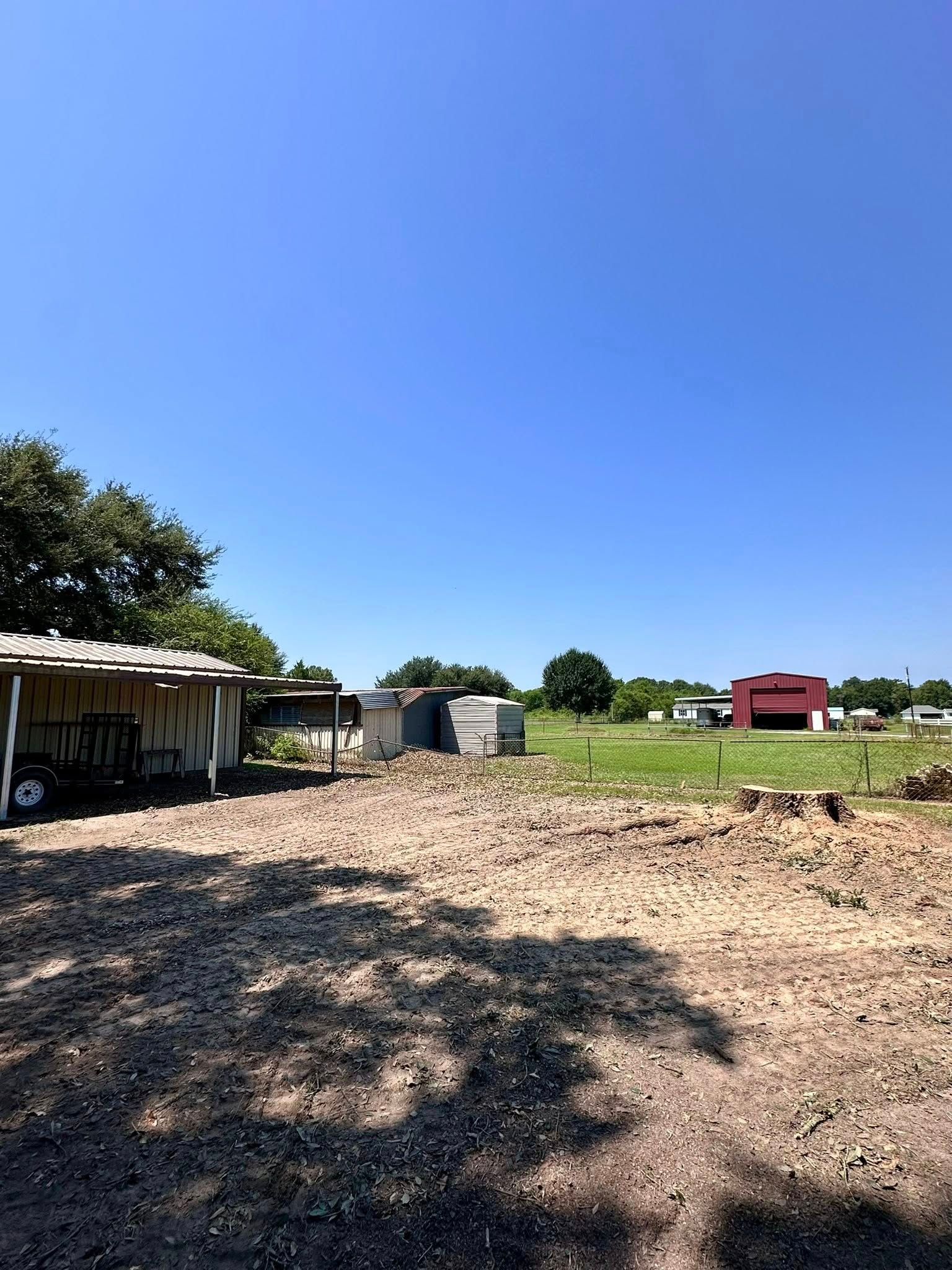 Open dirt lot with sheds, trees, and a red building under a clear blue sky