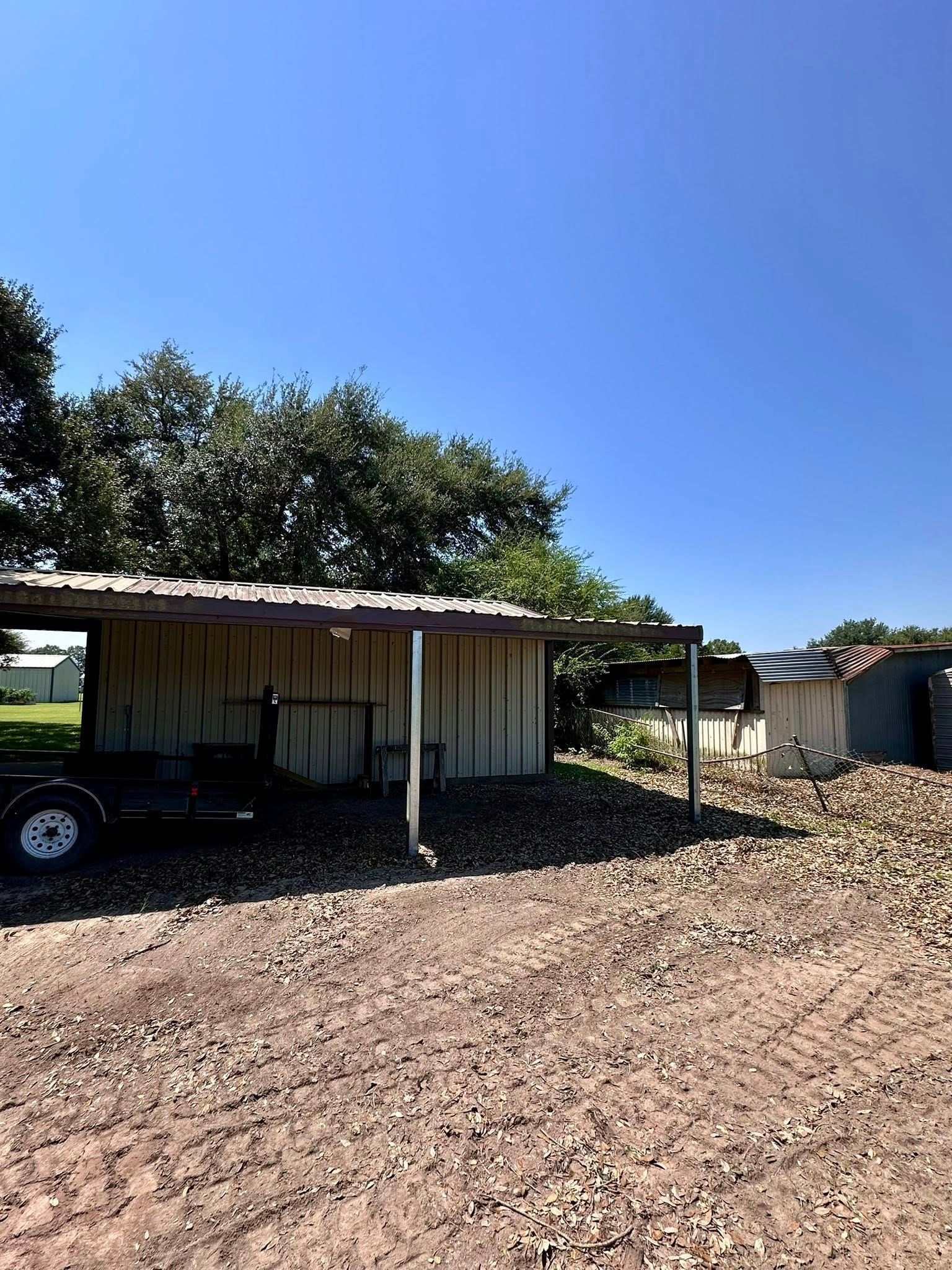 Rural carport with parked black vehicle, dirt ground, and trees under a clear blue sky