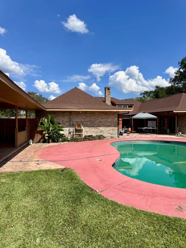 Backyard pool with pink deck, green lawn, and a stone house under a blue sky with clouds
