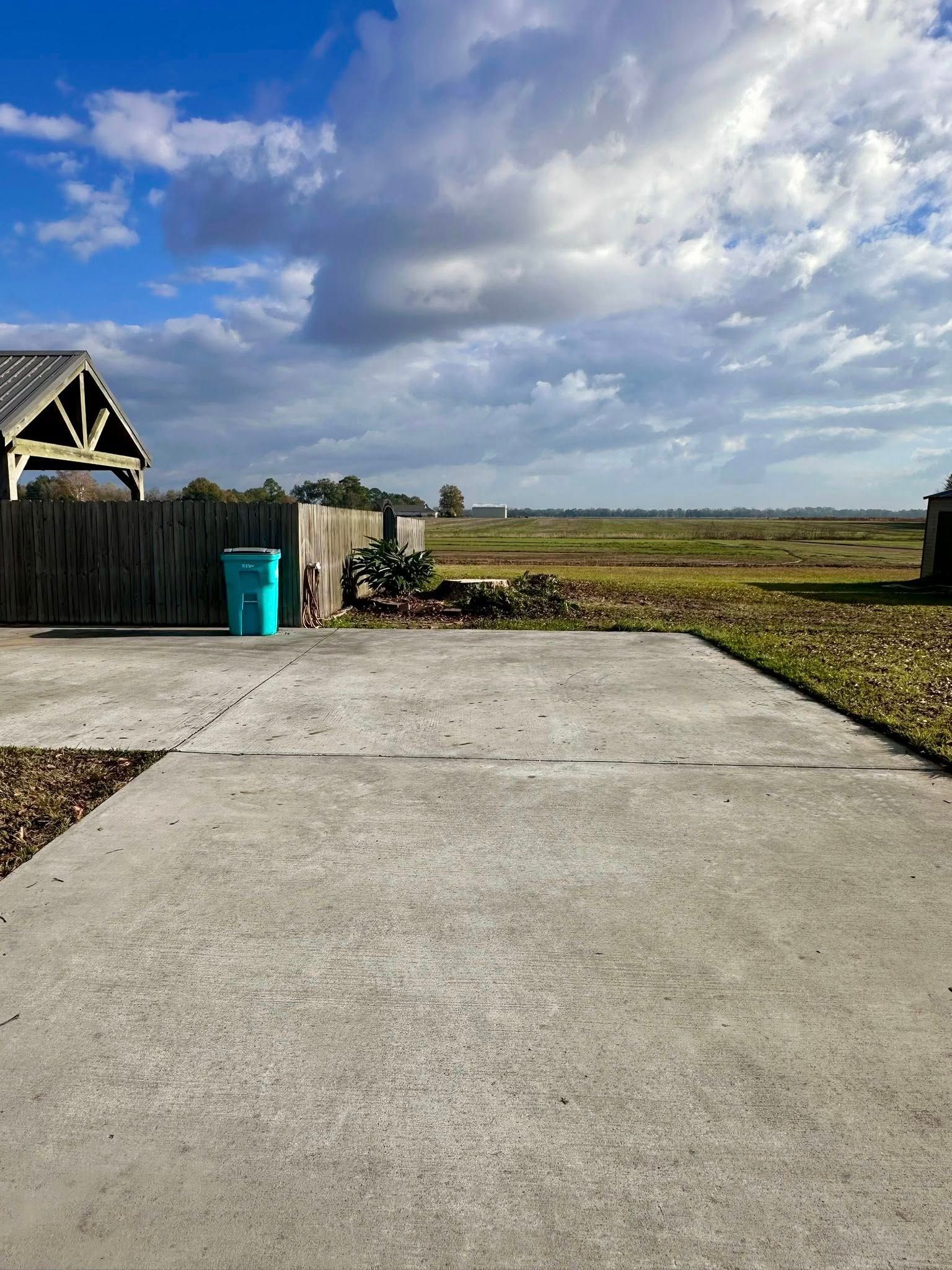 Concrete driveway leading to a grassy open field under a cloudy blue sky, with a fence and blue bin on the left.