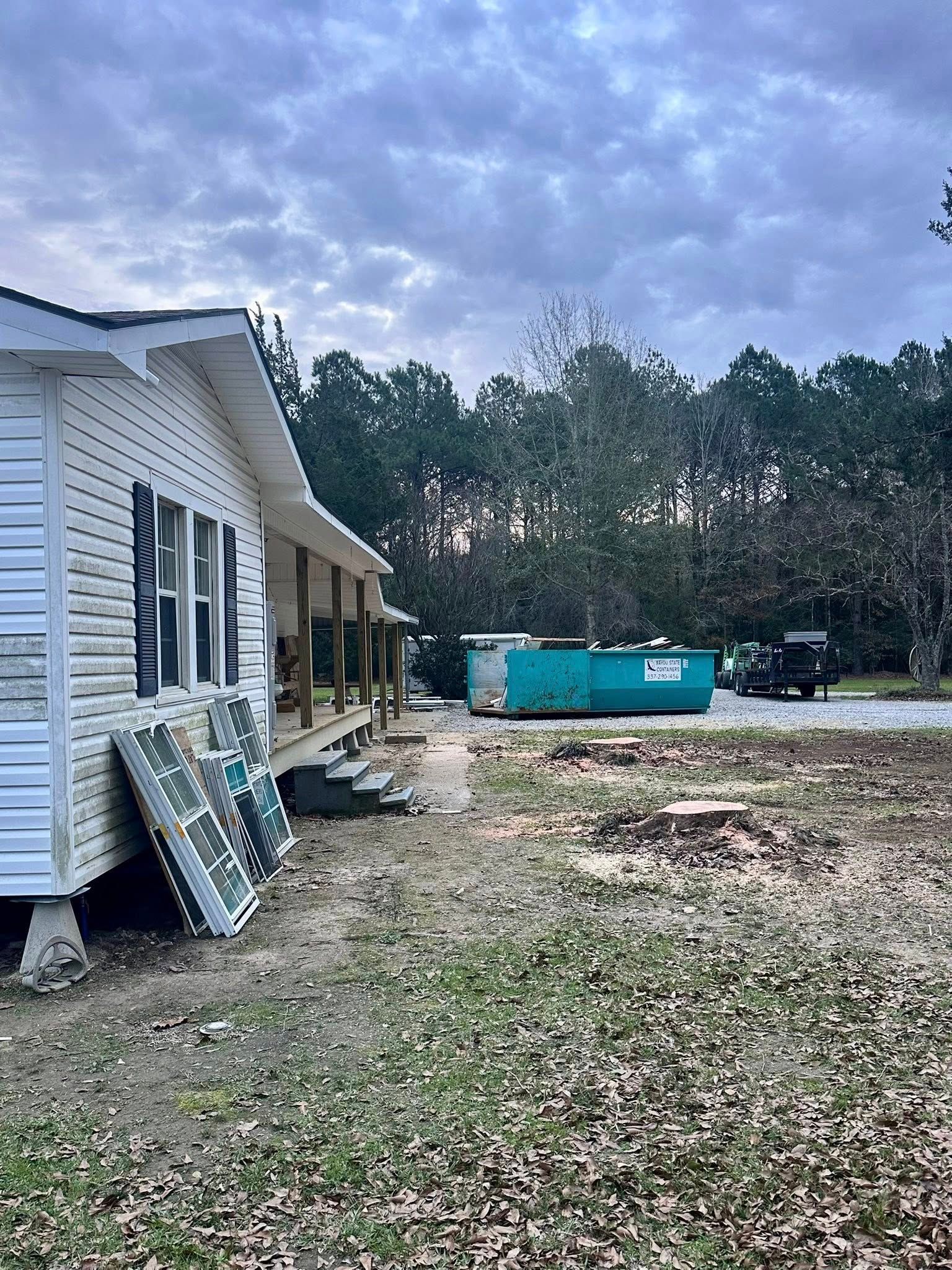 Partially built white house beside a dirt yard, with a blue dumpster and trees under a cloudy sky