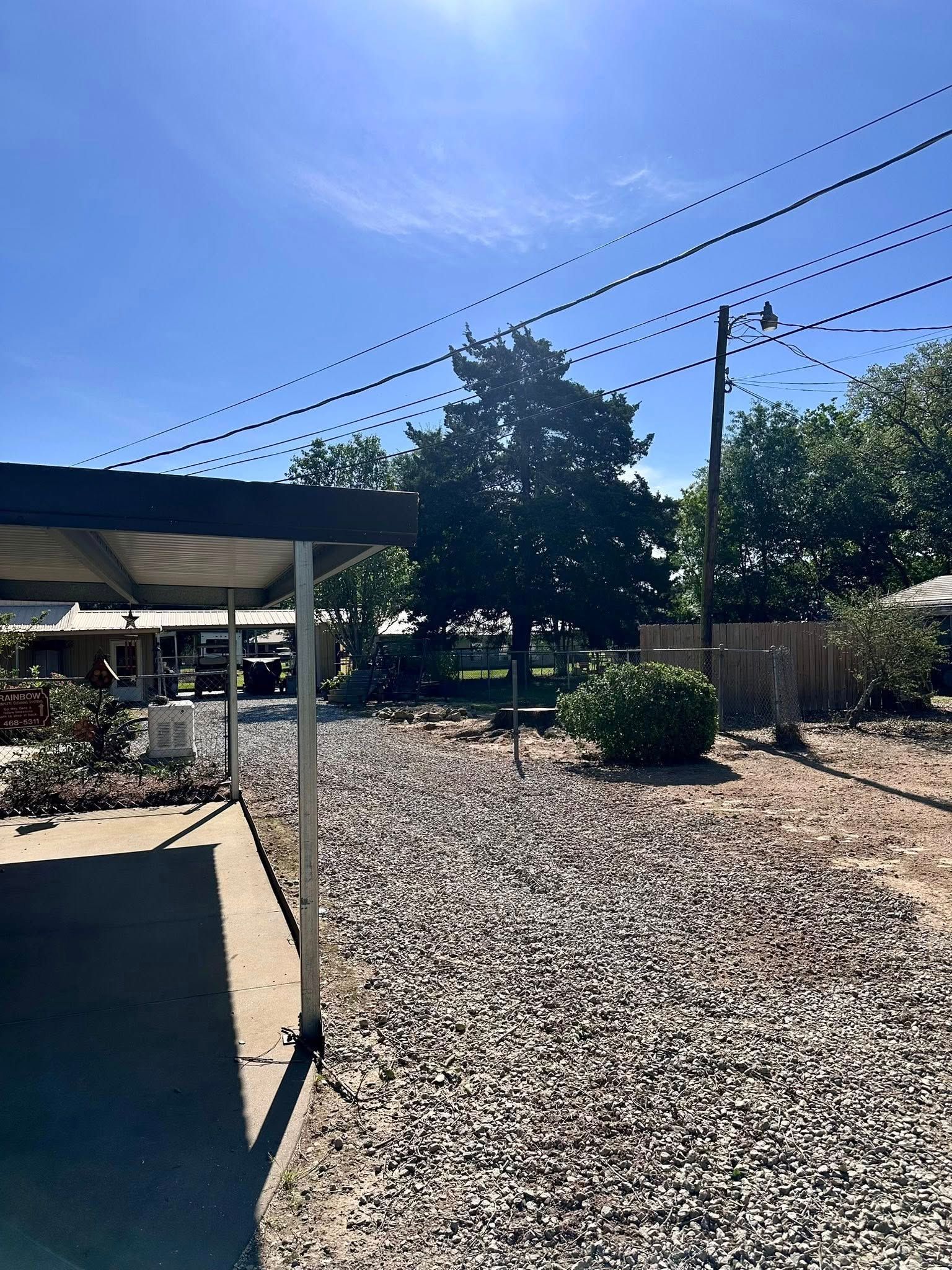 Sunny backyard with gravel ground, small trees, and a covered patio under power lines.