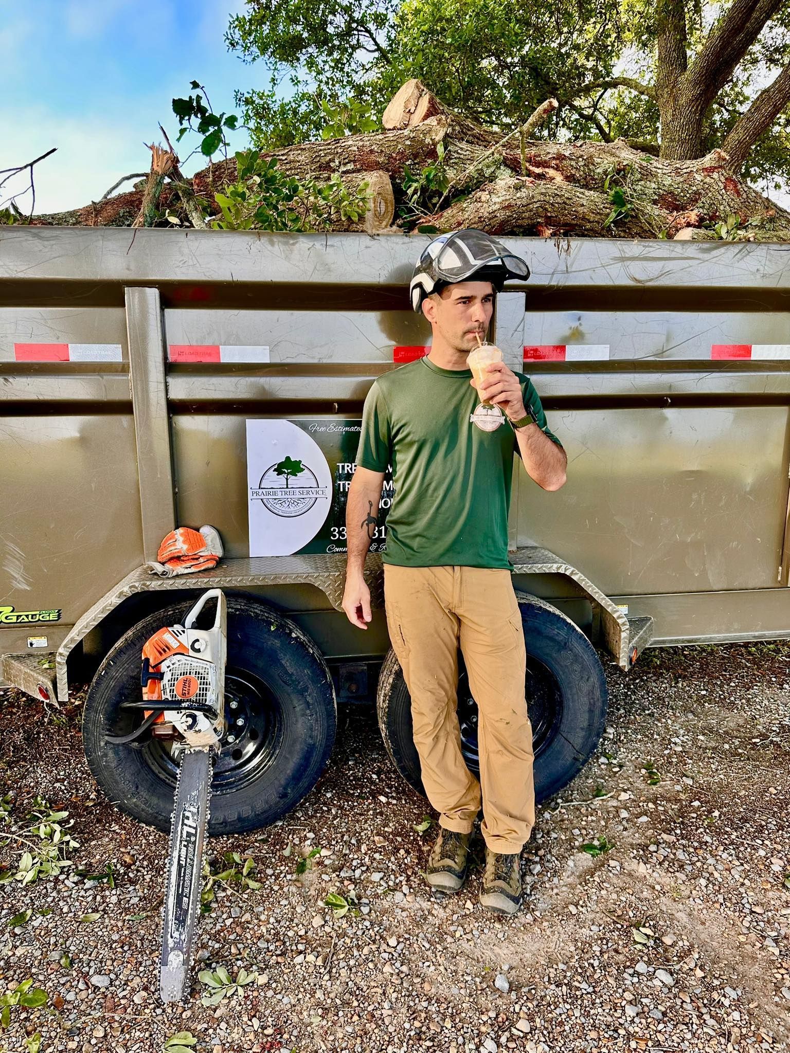 Person standing beside a small trailer loaded with branches, holding a drink outdoors.