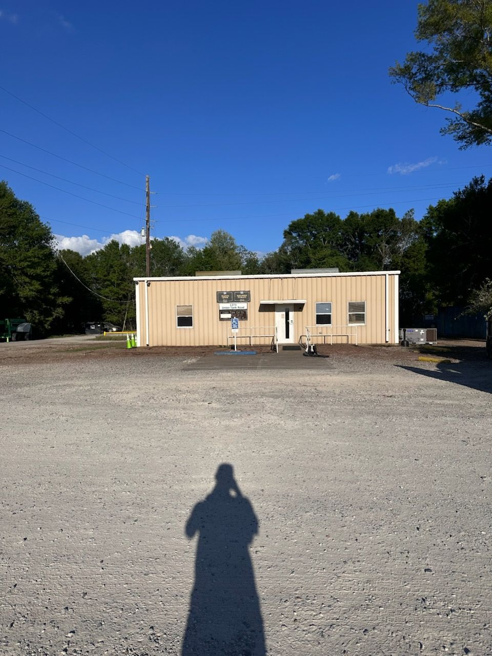 Small beige building in a gravel lot under a clear blue sky, with trees in the background and a person’s shadow in front.