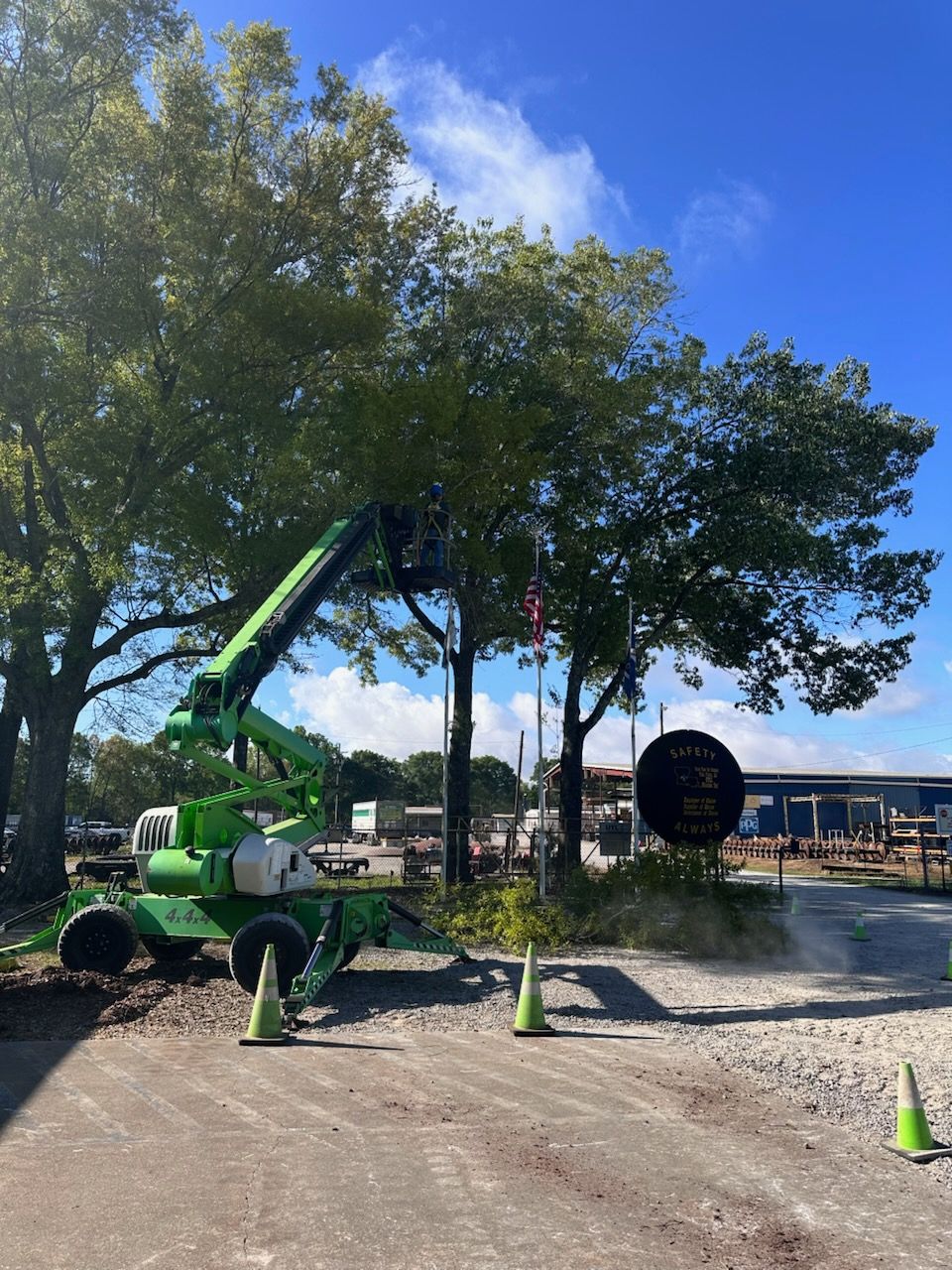 Green utility truck with raised crane parked on a gravel road under trees near a building.
