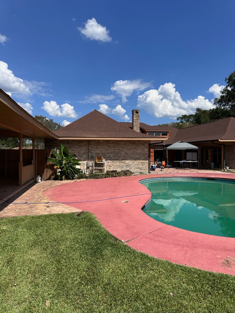 Backyard with swimming pool, red patio, lawn, and brick house under a blue sky